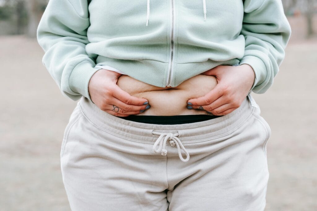 Crop anonymous plus sized female in casual hoodie touching and showing fat on belly while standing in park